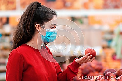Shopping. Portrait of a young woman in a medical mask on her face choosing apples in a supermarket. The concept of consumerism and Stock Photo