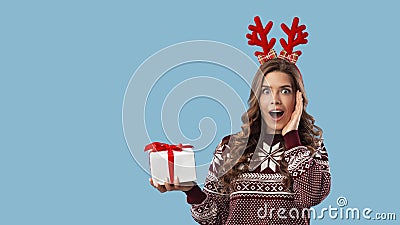 Shopping for Xmas. Surprised young lady in knitted sweater showing gift box on blue background, space for text Stock Photo
