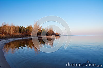 The shore of the reservoir. Sandy beach, autumn forest in the rays of the setting sun, calm expanse of water without waves Stock Photo