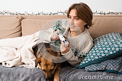 Short haired blond girl playing new video game on her console lying on couch with her dog, being immersed, happy, virtual reality Stock Photo