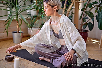 Short-haired woman set fire on stick of Palo Santo tree, lights a stick, engaged in yoga. Stock Photo