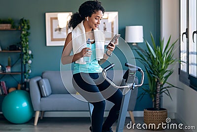 Afro young fitness girl using mobile phone while training on exercise bike at home Stock Photo