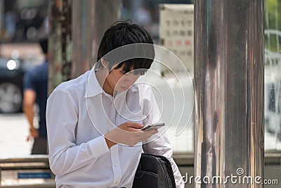 Shot of an attractive Japanese young man using his cellphone while waiting at bus stop. Editorial Stock Photo