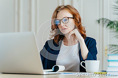 Shot of atttactive businesswoman concentrated in monitor of laptop computer, has serious focused gaze, wears optical glasses for Stock Photo