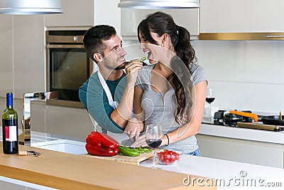 Beautiful young couple having romantic moments, man feeding her wife with green pepper and smiling in the kitchen at home Stock Photo