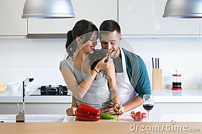 Beautiful young couple having romantic moments, woman feeding her husband with green pepper and smiling in the kitchen. Stock Photo