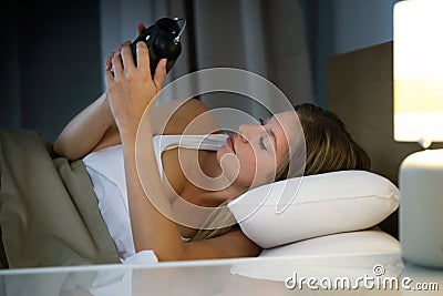 Beautiful young exhausted woman suffering insomnia holding and looking clock in bedroom at home. Stock Photo