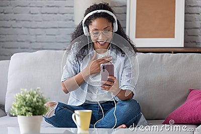 Beautiful young woman having video conversation with friends with headphones while using her smartphone sitting on sofa at home Stock Photo