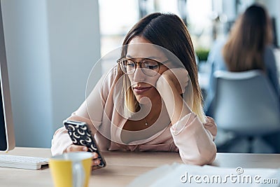 Boring young business woman sending messages with mobile phone while sitting in modern startup office Stock Photo