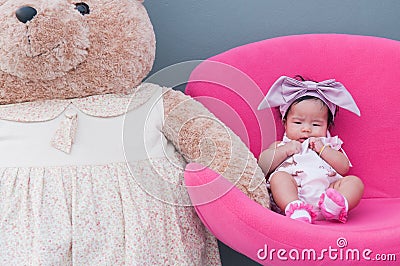 A shot of a cute baby girl with purple headband and big teddy bear while sleeping and playing on the pink chair / Focus at infant Stock Photo