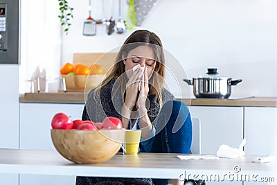 Illness young woman sneezing in a tissue while sitting in the kitchen at home Stock Photo