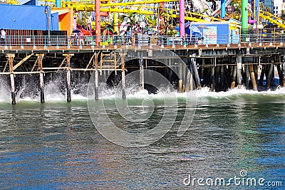 A shot of lush green ocean water and waves crashing into the legs of a wooden pier with colorful carnival rides on top of the pier Editorial Stock Photo