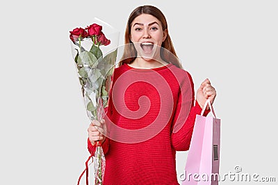 Shot of pleased overjoyed woman with dark hair, dressed in oversized red sweater, carries roses and gift bag, recieves package, Stock Photo