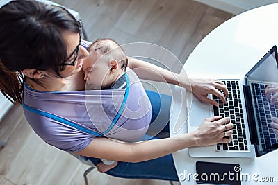 Pretty young mother with her baby in sling working with laptop at home Stock Photo