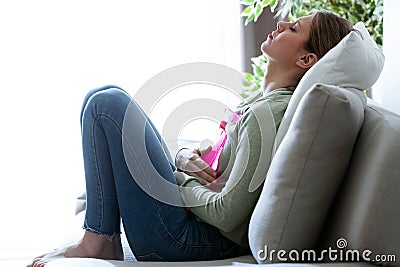 Unhealthy young woman with stomachache using a hot water bag while sitting on the couch at home. Stock Photo