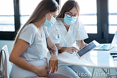 Young beautiful gynecologist wearing a hygienic face mask while showing to pregnant woman ultrasound scan baby with digital tablet Stock Photo