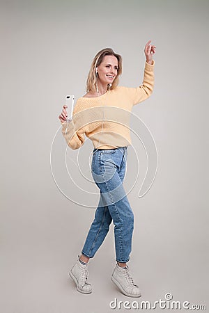 Shot of young blonde woman dancing while holding her smartphone in tased hand and listening to the music in earphones and smiling Stock Photo