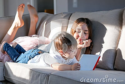 Siblings reading book together on couch with feet up. Hard light. Sister and her little brother spending time together at home rea Stock Photo