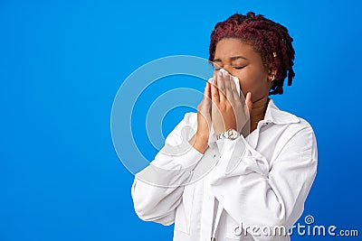 Sick afro woman sneezing in paper napkin against blue background Stock Photo