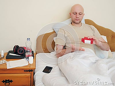 Sick bald unshaven man in his bad with medicine pills in his hands in his bed, concept flue and corona virus treatment Stock Photo