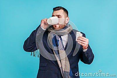 Sick businessman wearing official style suit and wrapped in scarf, holding pills in hands and drinking warm tea, suffering from Stock Photo