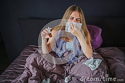 Sick and ill young woman sits on ned and sneezing in white napkin. She holds nose spray in right hand. Young woman Stock Photo