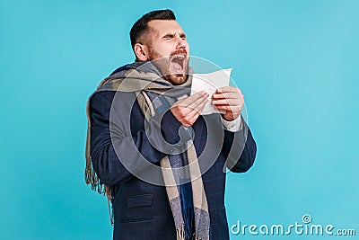 Sick man in official style suit and wrapped in scarf sneezing tissue, feeling unwell with runny nose, suffering allergy or Stock Photo