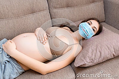 Sick pregnant woman is lying on the couch at home. Protect your health with protective medical mask. infection. Global Stock Photo