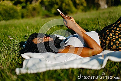 Side portrait of the young attractive african girl with green eye shadows laying on the grass and browsing and chatting Stock Photo
