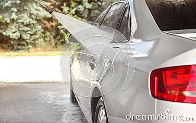 Side of silver car washed in self serve carwash, jet water spraying on metal, blurred sun lit trees background Stock Photo