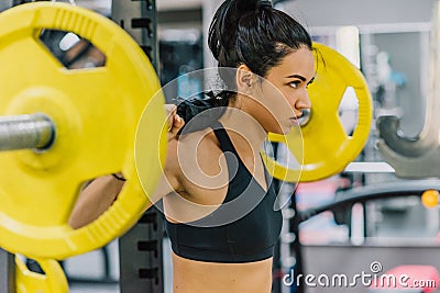 Side view of beautiful young woman doing workout in the gym, pushing yellow barbell on shoulders. Female hard workout for cross Stock Photo