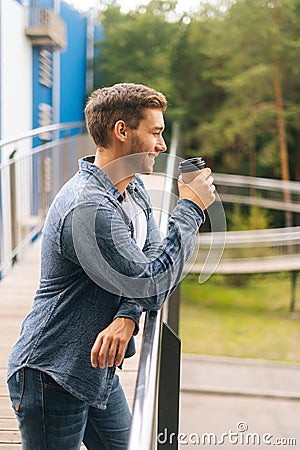 Side view of confident thoughtful young man standing on balcony terrace and drinking coffee leaning on railing of office Stock Photo