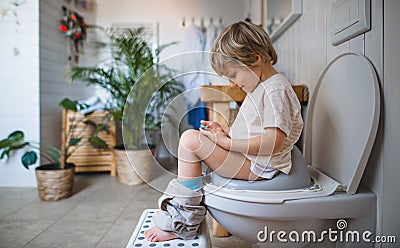 Side view of cute small boy sitting on toilet indoors at home, using smartphone. Stock Photo