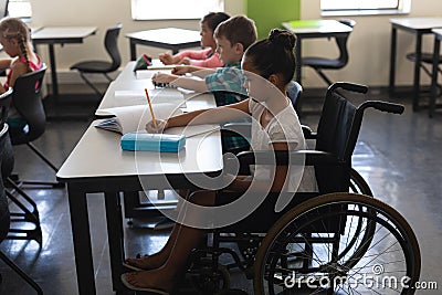 Side view of disabled schoolgirl with classmates studying and sitting at desk in classroom Stock Photo