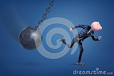 A side view of a fit businessman with a penny bank instead of his head running from a demolition ball. Stock Photo
