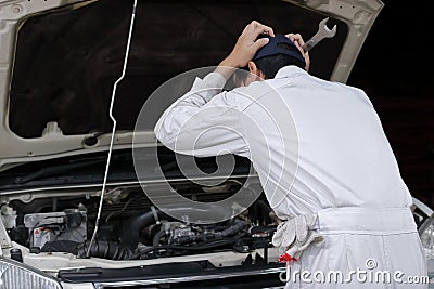 Side view of frustrated stressed young mechanic man in white uniform touching his head with hands against car in open hood at the Stock Photo