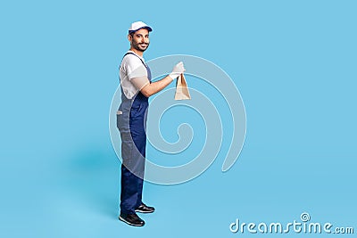 Side view, happy courier in blue uniform holding groceries bag and smiling to camera, carrying parcel Stock Photo