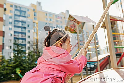 Side view image of happy pretty little girl climbing on a rope at playground outdoor. Cute preschool toddler girl playing outside Stock Photo