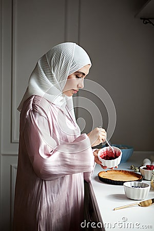 Side view portrait Muslim woman cooks dessert cake at kitchen, arabian young model in hijab and abaya. Islamic Stock Photo