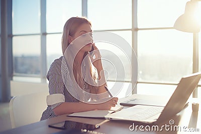 Side view portrait of young businesswoman having business call in office, her workplace, writing down some information Stock Photo
