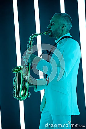 Side view saxophone player in a white suit against the backdrop of neon lamps with blue backlight. Saxophonist jazzman Stock Photo