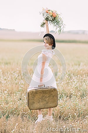 The side view of the smiling bride carrying the vintage suitcase, keeping up the colourful bouquet and looking at the Stock Photo