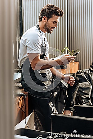 Side view of smiling young hairstylist in apron using smartphone Stock Photo