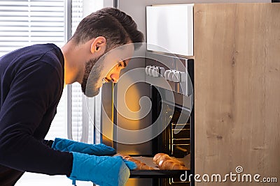 Smiling Young Man Taking Out Tray Of Croissants From Oven Stock Photo