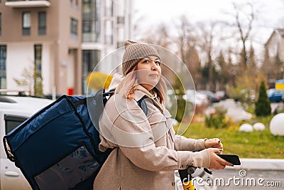 Side view of tired food delivery woman with thermo backpack holding mobile phone waiting next order standing with Stock Photo