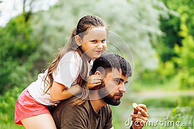 Side view of wonderful family sitting on picnic in park forest around trees. Little daughter sitting on fathers back. Stock Photo