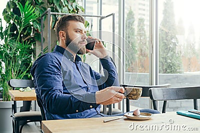 Side view.Young bearded attractive businessman in blue shirt is sitting at wooden table near window in restaurant Stock Photo