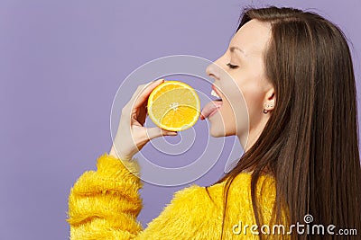 Side view of young woman in fur sweater hold licking half of fresh ripe orange fruit isolated on violet pastel Stock Photo