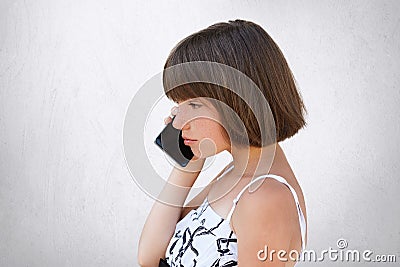 Sideways portrait of little girl with bobbed hair, wearing white dress, speaking over cell phone with serious expression. Stylish Stock Photo