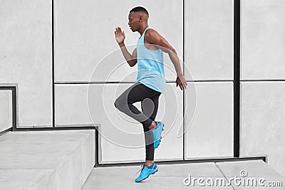 Sideways shot of determined Afro American sportsman runs up stairs, has goal to overcome breath shortage, wears vest Stock Photo